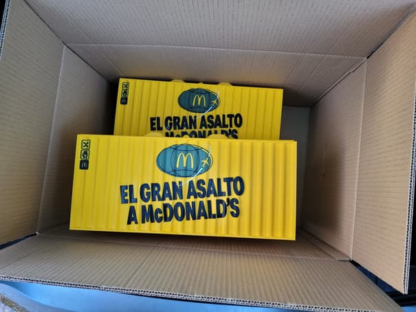 Dos cofres amarillos con forma de contenedor marítimo rotulados con la marca McDonald’s, dentro de una caja de cartón
